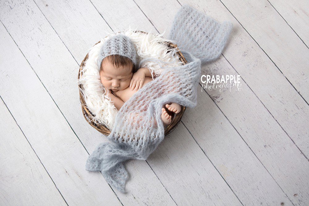 White and light blue new baby photo. Baby boy sleeping curled up in wooden nest bowl with white fur.