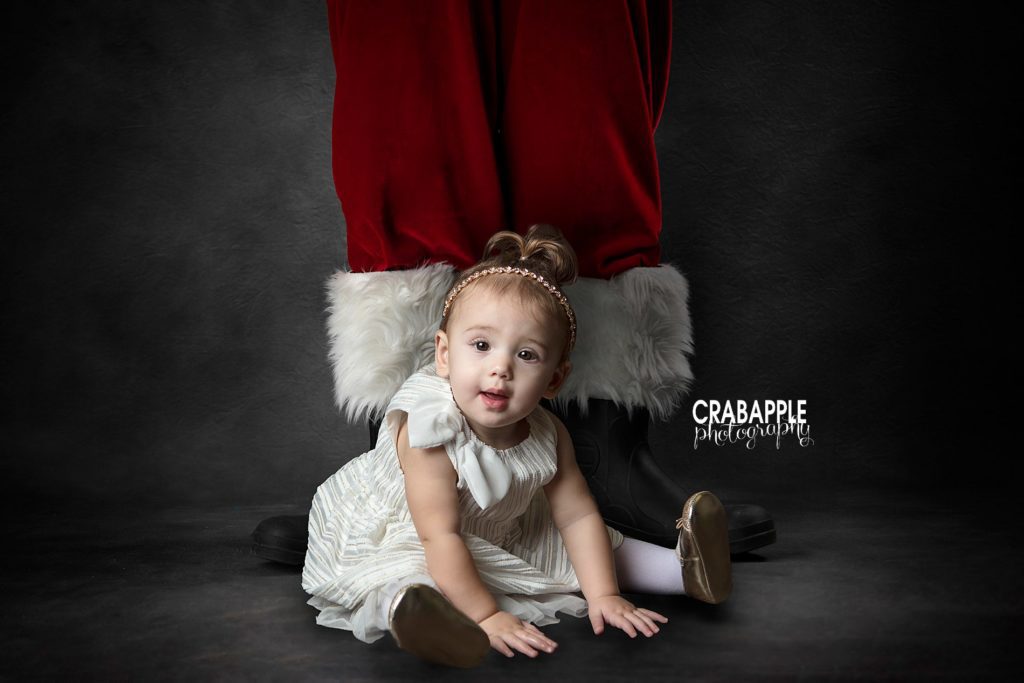 Photo of a baby girl sitting on the floor while Santa stands behind her. You can only see Santa's legs including the classic red velvet pants with white fur trim and black boots. This is a digital composite Santa Claus photo in Andover MA.