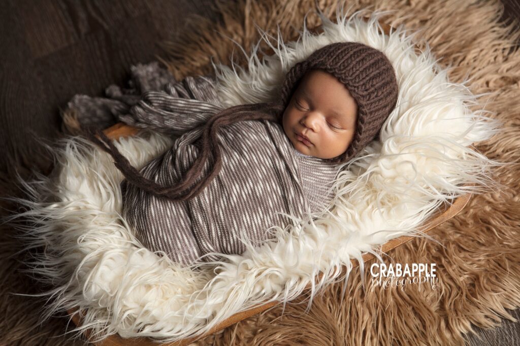 Brown and white newborn photo styling inspiration with a baby boy swaddled in brown and white stripes, wearing a dark brown knit hat. He is on a white fur that is placed o