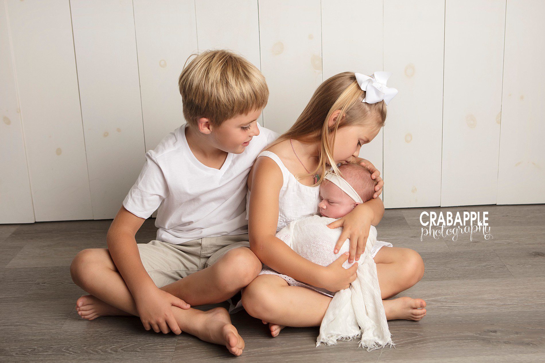 Two siblings in white pose with their newborn baby sister
