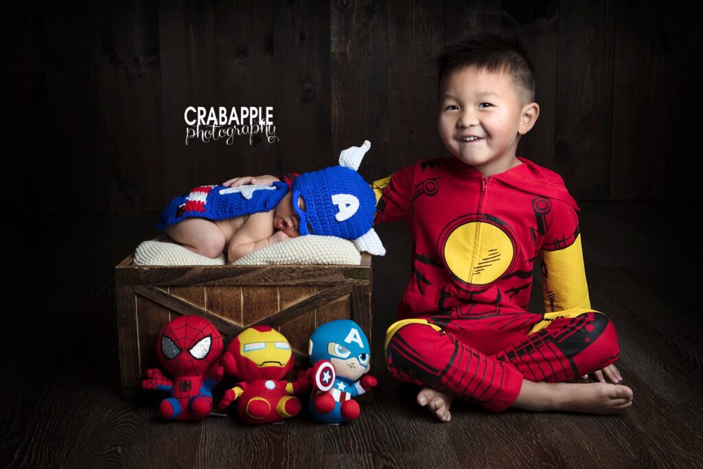 Marvel Avengers themed newborn photos. Baby boy is wearing a knit Captain America costume and sleeping on a wooden crate. Big brother is wearing an Iron Man costume. Three stuffed toys of Captain America, Iron Man, and Spider-Man sit in front.