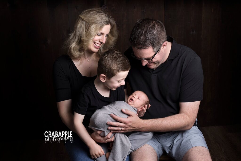Family photo of mom, dad, and 4 year old brother all wearing black tee shirts. Big brother is holding newborn brother swaddled in gray.