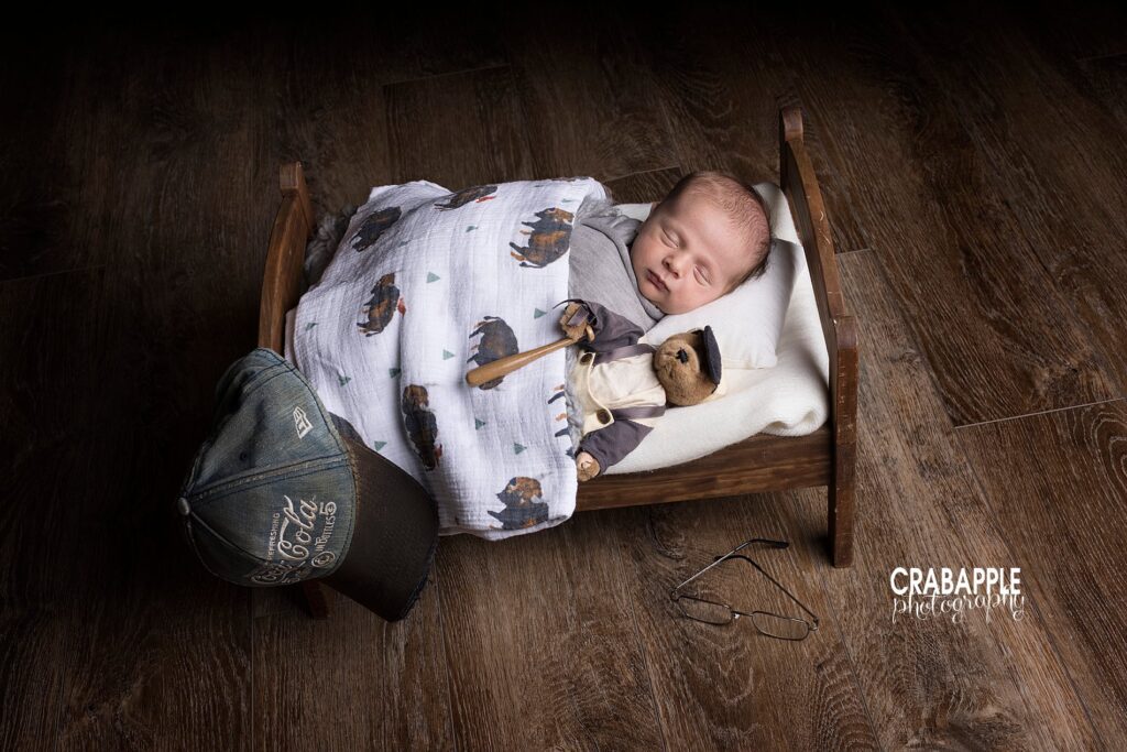 Newborn baby boy sleeps in a tiny wooden bed. He has a vintage teddy bear holding a baseball bat, a baseball hat, and reading glasses around him in honor of a lost loved one.