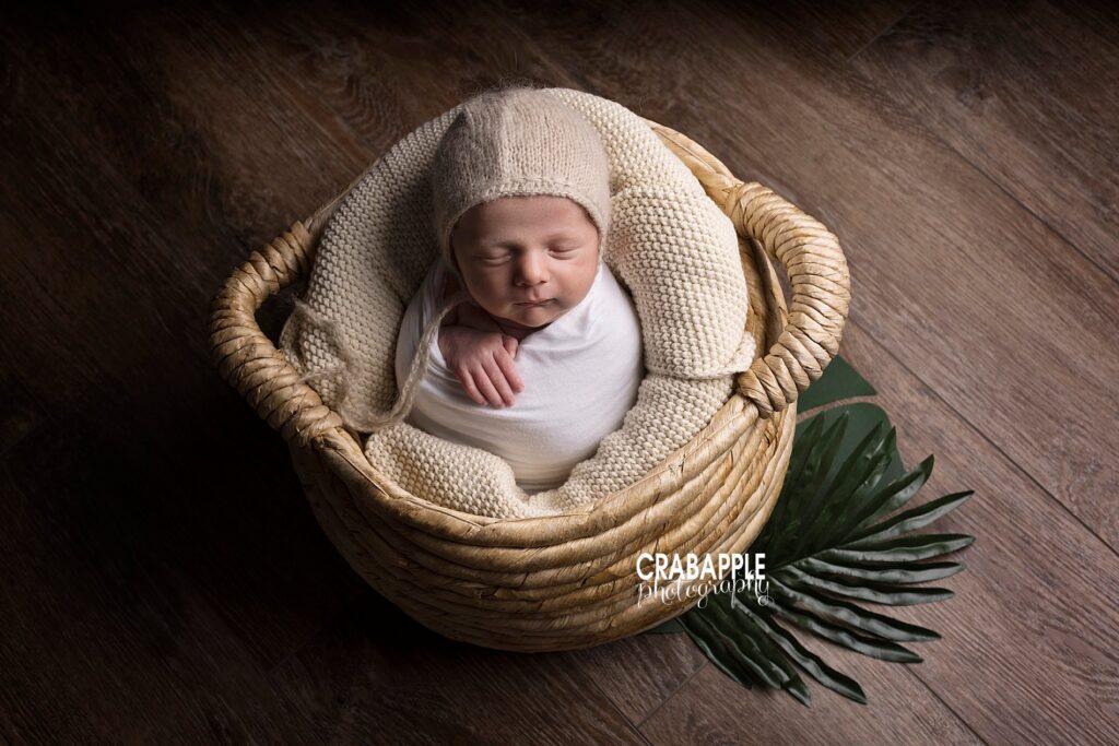 Classic newborn baby boy photo of sleeping baby wrapped in white inside a tan basket. There are also faux tropical leaves.