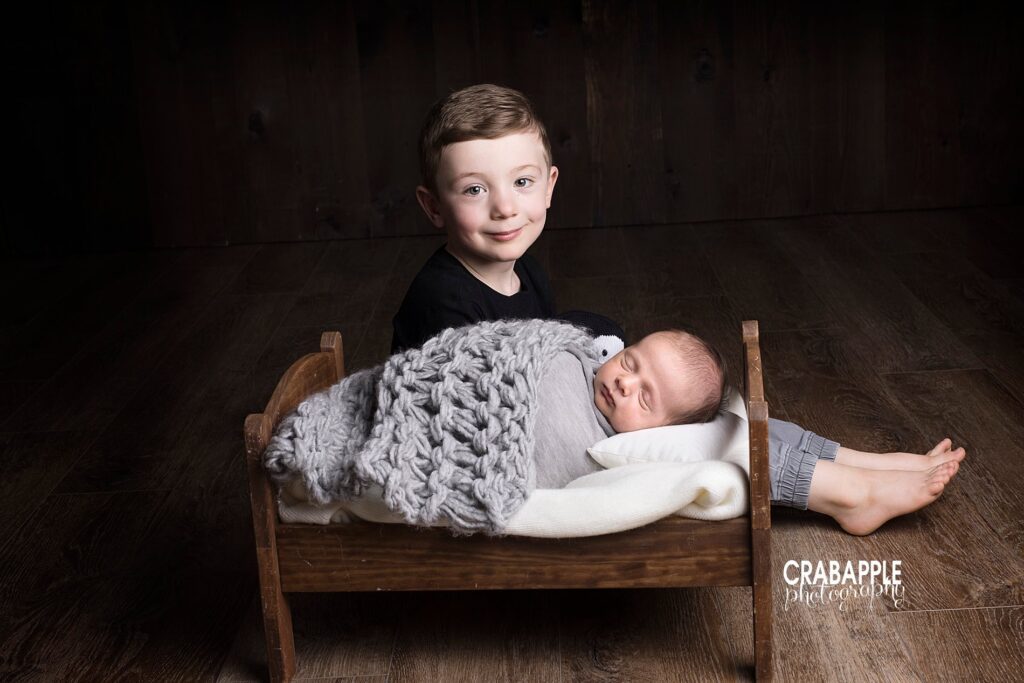 Photo of two brothers, 4 year old big brother and his newborn baby brother. Newborn is in a wooden bed swaddled in gray while big brother sits behind the bed smiling at the camera.