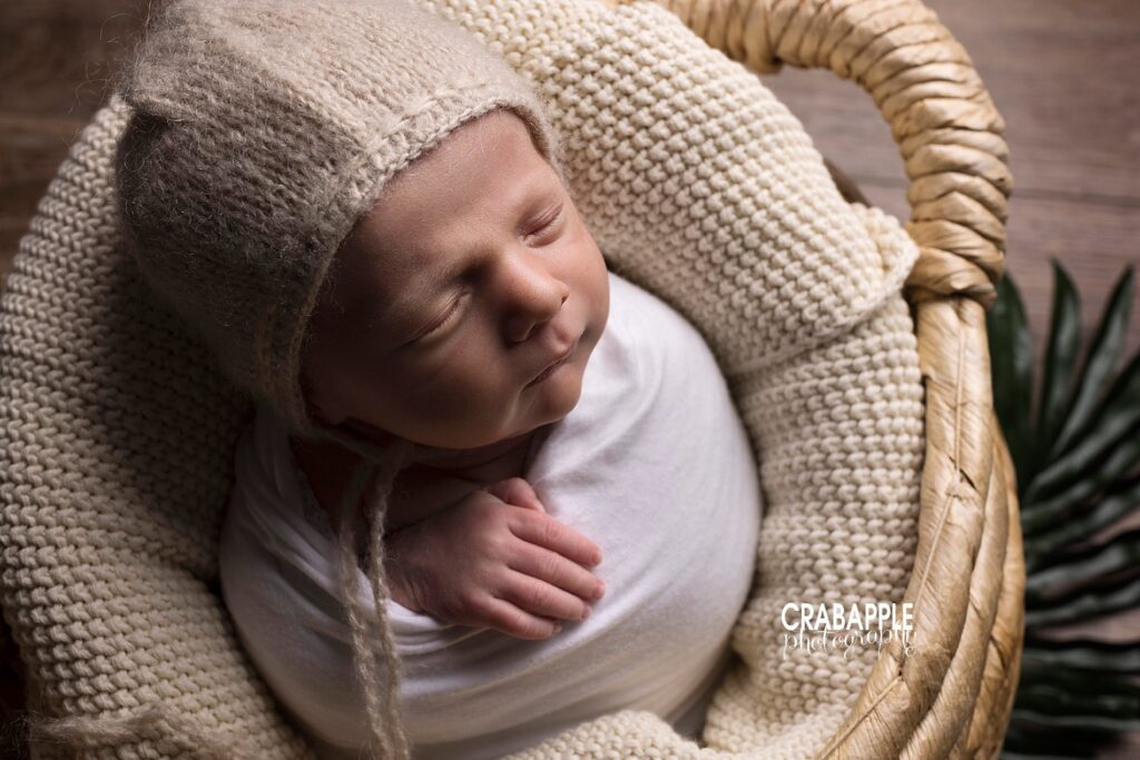 Close up photo of a newborn boy wrapped in white wearing a cream bonnet. He is in a rattan basket with a cream blanket.