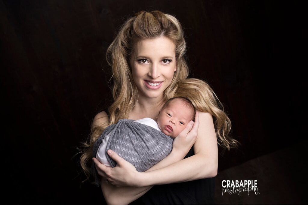 Photo of mom holding her newborn baby boy in her arms. Baby boy's eyes are slightly open and they're both looking at the camera in front of a dark background.