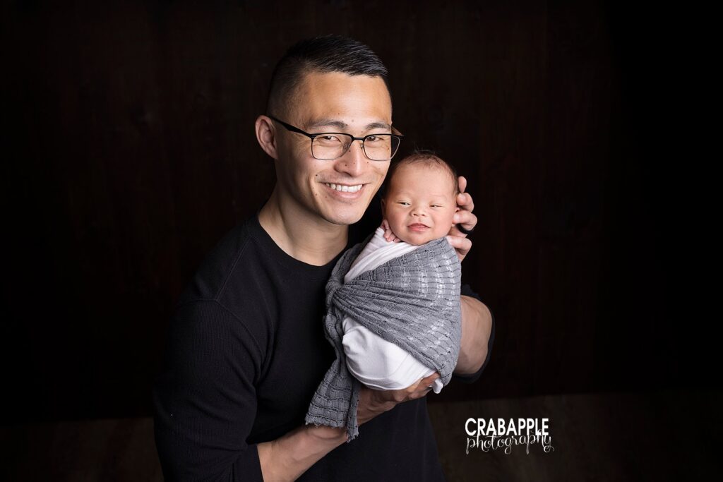 Photo of dad holding her newborn baby boy in his arms. Baby boy's eyes are slightly open and they're both looking at the camera in front of a dark background.