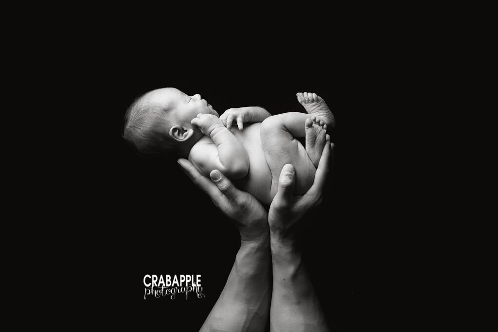 Black and white portrait of dad's hands holding newborn baby boy in front of a fully black background. 