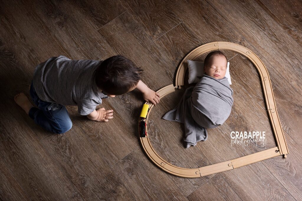 Sibling and newborn photo ideas where newborn baby boy is laying on the floor swaddled with train tracks in the shape of a heart around him. Toddler big brother plays with the train on the tracks.