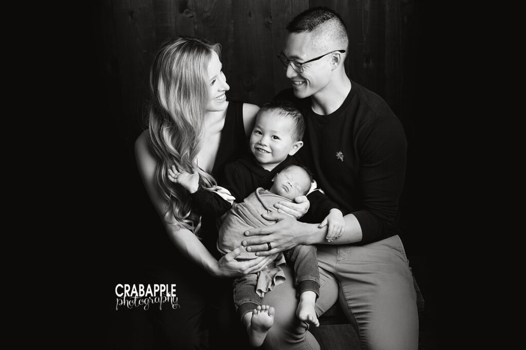 Black and white family portraits. Mom and dad look at each other and smile while toddler brother smiles at the camera and newborn brother sleeps swaddled in their arms.