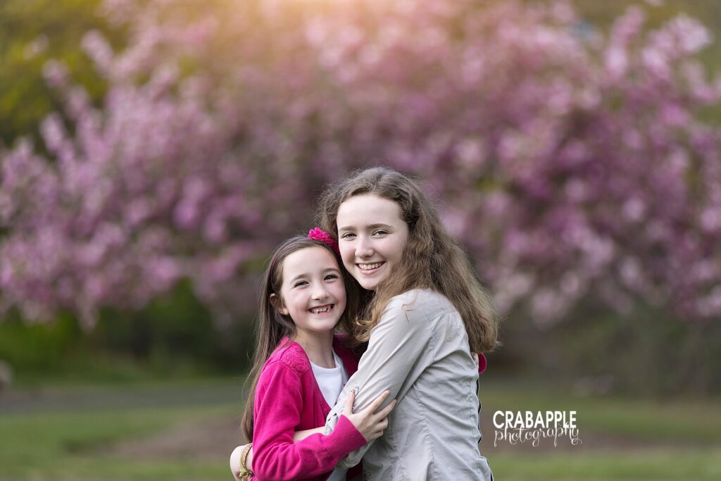 Portrait of two sisters, ages 14 and 8, hugging outdoors in front of a pink flowering tree.