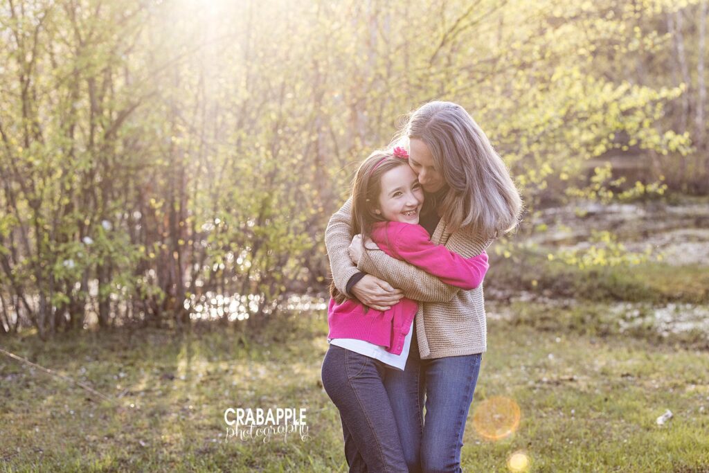 Mother and 8 year old daughter standing outside, daughter is smiling at the camera while mom gives her a big hug.