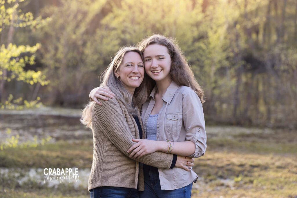 Family portrait of mom and teenage daughter, standing outside with arms around one another and smiling at the camera.
