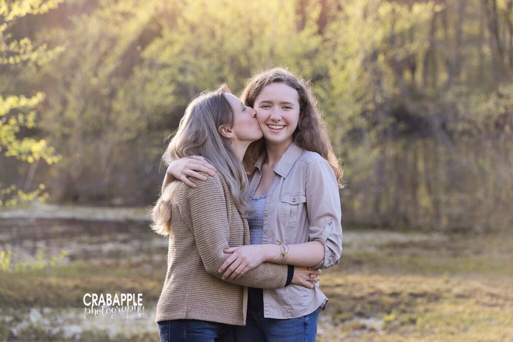 Family portrait of mom and teenage daughter, standing outside with arms around one another. Daughter is looking at the camera while mom kisses her cheek.