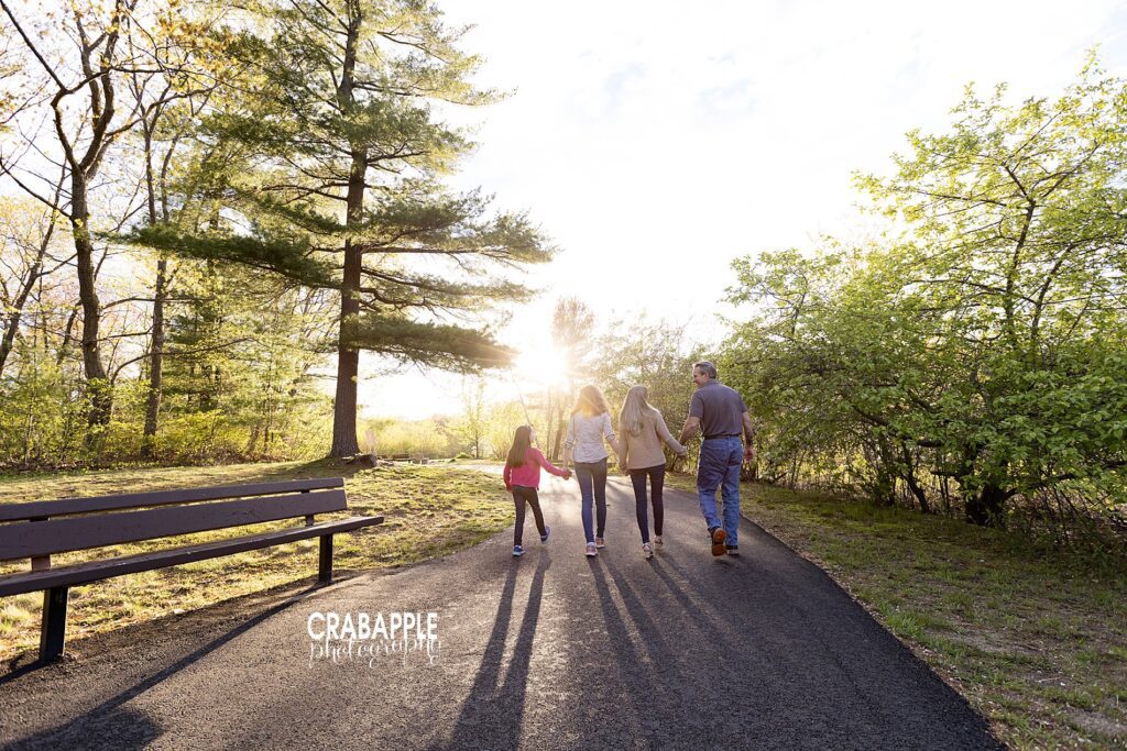 Portrait of a family of four walking down a tree lined paved walkway outside, away from the camera. The glow of the sun is seen overhead.