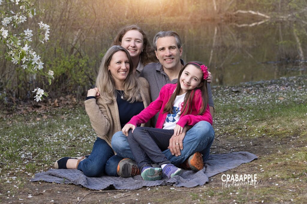 Photo of a family of four sitting on a gray blanket outdoors. There is a small body of water and a flowering tree behind them.
