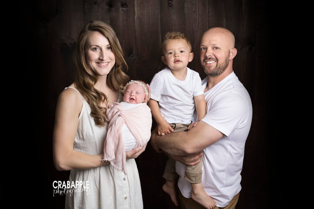 Simple and classic family photos with newborn baby, toddler brother, and parents in front of a black backdrop.
