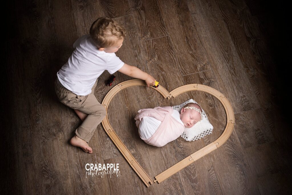 Sibling and newborn photo ideas, newborn baby girl is on the floor with wooden train tracks shaped in a heart around her. Toddler brother is playing with a train on the tracks.