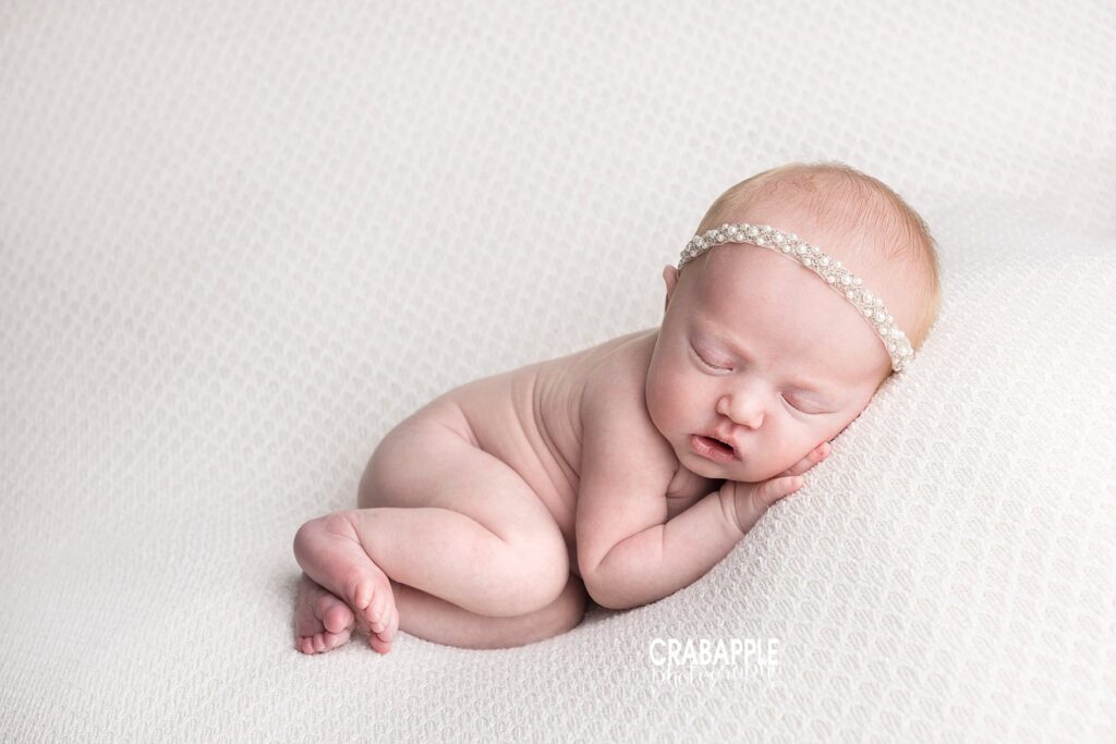 Clean, simple, and serene newborn photo of a baby girl laying on her side on a white blanket wearing a dainty pearl headband.
