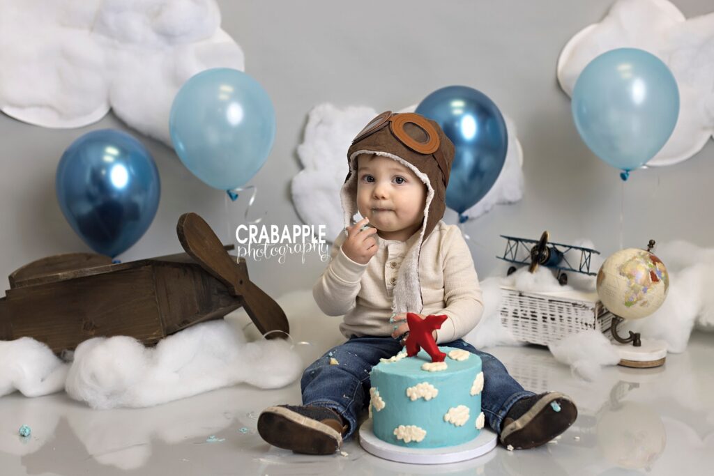 Airplane themed cake smash. One year old boy sits in front of a gray backdrop adorned with fluffy cotton clouds and blue balloons. Two vintage style airplanes are behind him, and a vintage globe. He wears a pilot's hat and has a light blue cake with white clouds and a red airplane cake topper.