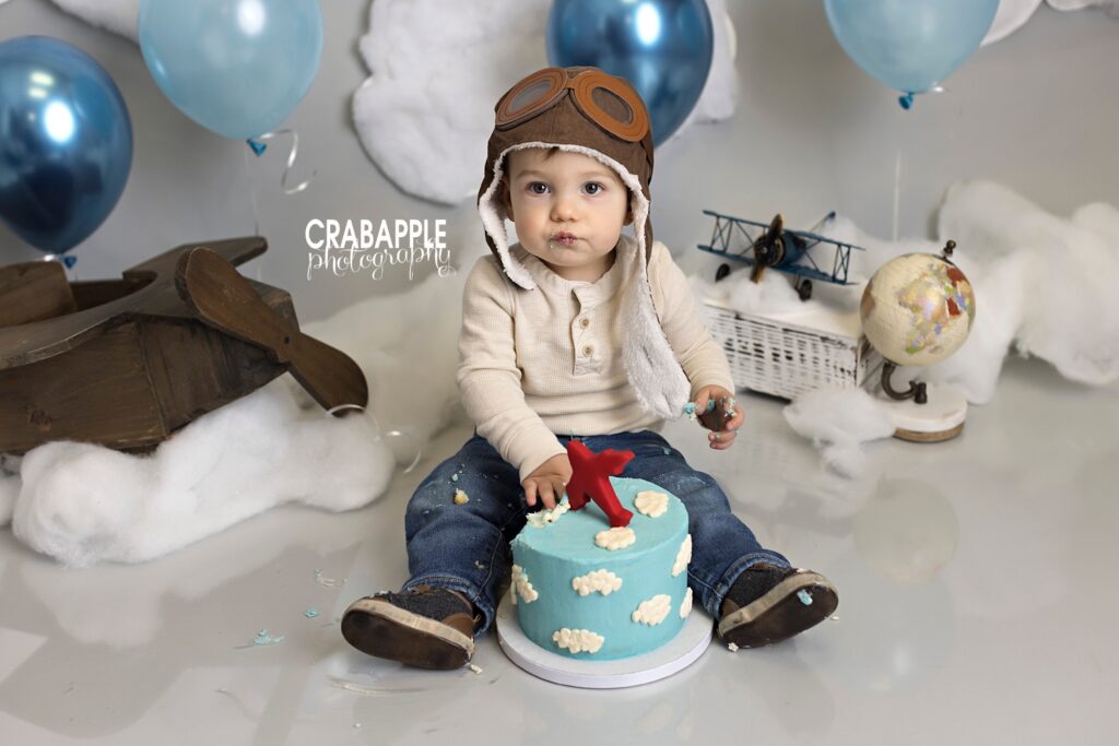 Airplane themed cake smash photos. 1 year old boy wearing jeans, a cream top, and a pilot's hat sits eating a blue cake with white clouds and a red airplane. Behind him you can see vintage planes, a globe, blue balloons and cotton clouds.