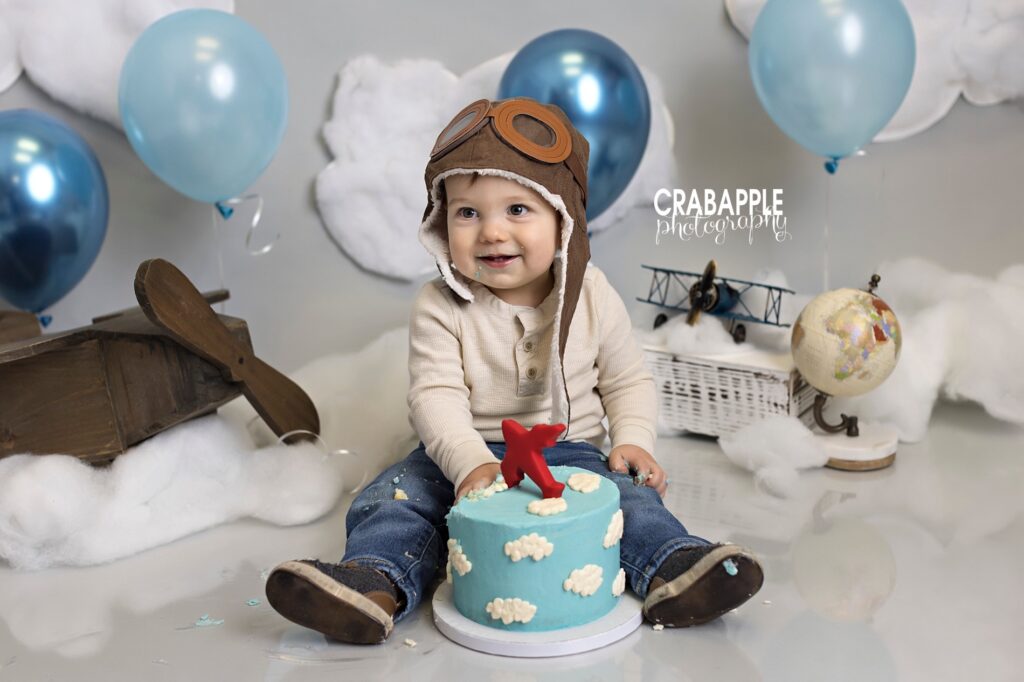 Smiling one year old boy looks away from the camera while grabbing his blue and white sky birthday cake topped with red airplane. He is on a gray backdrop. Behind him are vintage airplanes, globes, balloons, and cotton clouds.