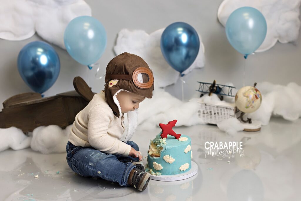 Side profile portrait of a one year old boy with his partially smashed airplane birthday cake. The set is completed with cotton clouds, blue balloons, vintage airplanes and a globe.