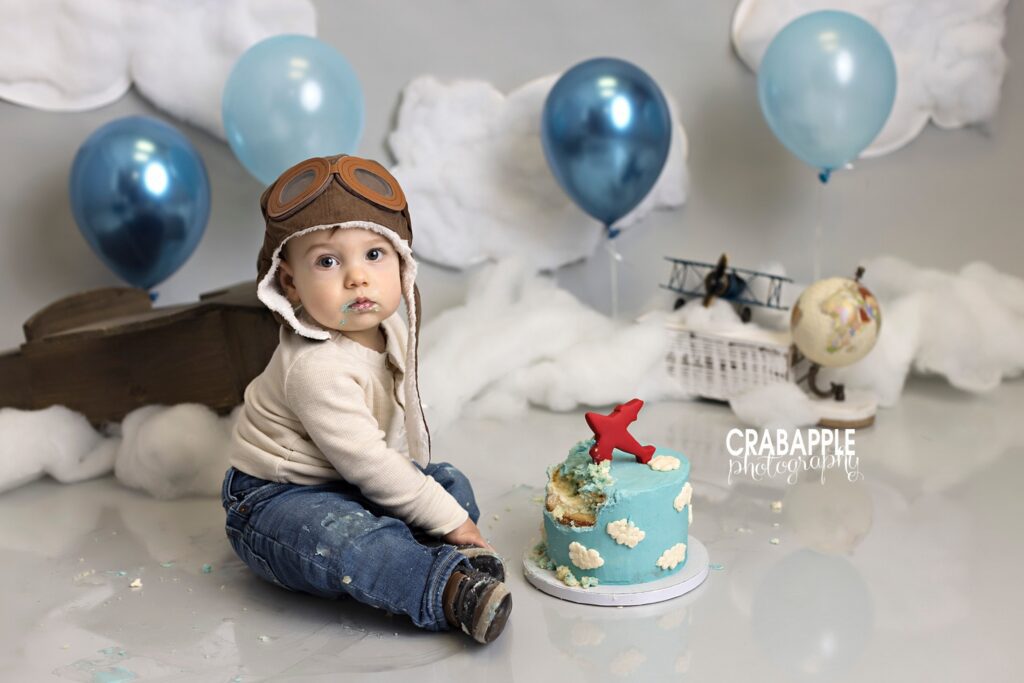 1 year old boy looks at the camera with blue frosting on his face from his blue and white sky birthday cake topped with red airplane. Behind him is a set featuring blue balloons, cotton clouds, and other props.