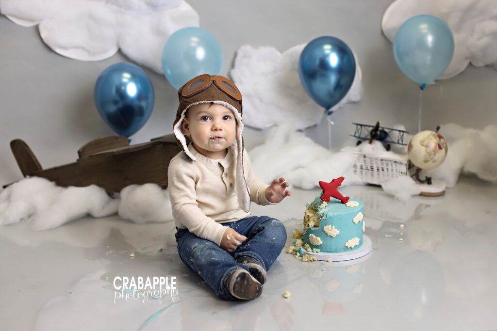 A cake smash set design featuring vintage airplane toys, a globe, cotton clouds, blue balloons. A one year old boy sits in front with a pilot hat, a sky themed cake, and frosting all over him.
