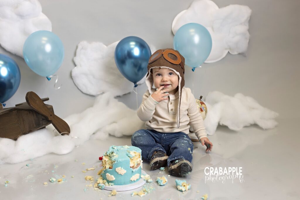 Sky themed cake smash photo. A gray backdrop is covered with cotton clouds and blue balloons. The birthday cake is blue with white clouds. Baby boy is wearing a pilot's hat.