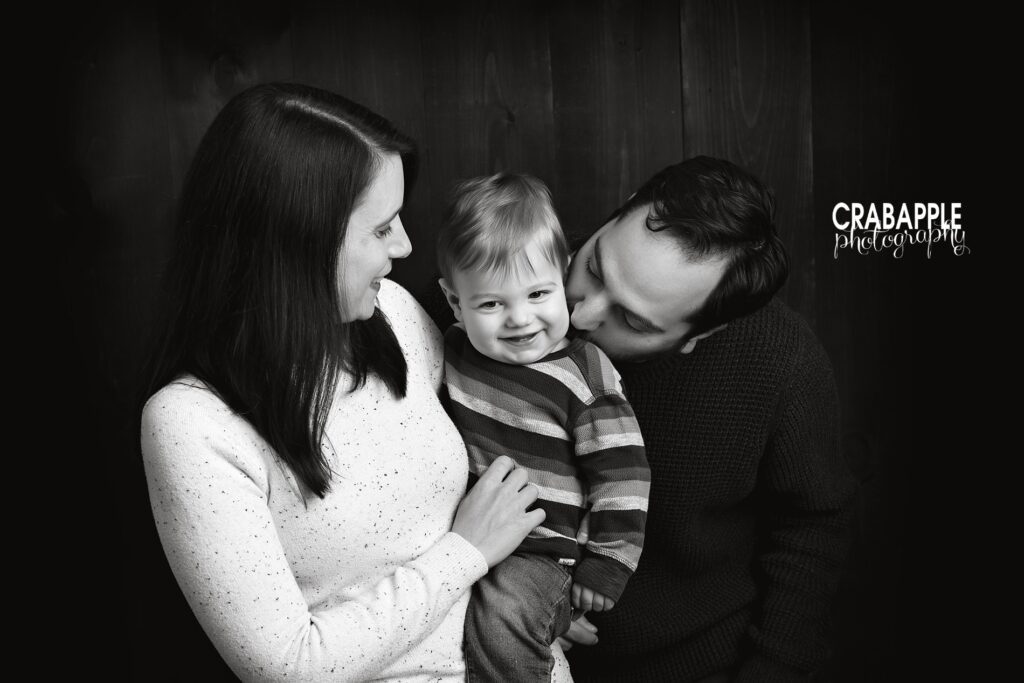 Black and white photo of mom, dad, and one year old son. The baby is smiling while his dad kisses him and mom looks at him smiling.