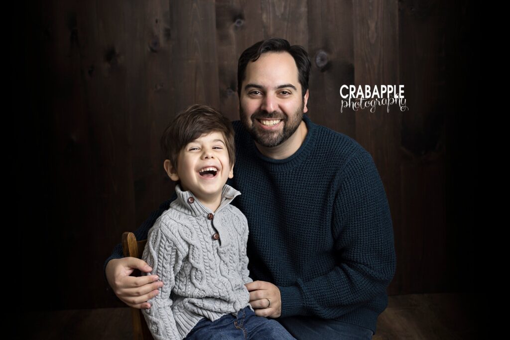 Photo of dad and his 5 year old son. Both are looking at the camera and smiling in front of a dark wood background.