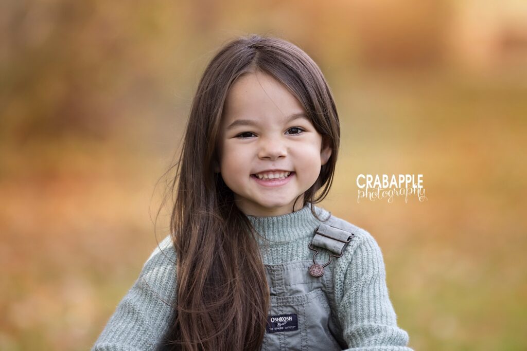 Portrait of a 6 year old girl in a gray knit sweater and gray overalls smiling at the camera.