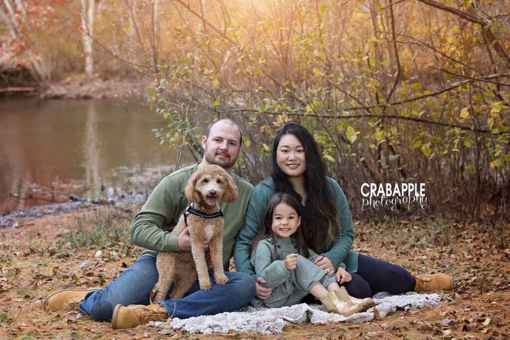 Family portraits outside during autumn in New England with autumn leaves on the ground. Mom, dad, 6 year old daughter and the family puppy all sit on the ground on a blanket.