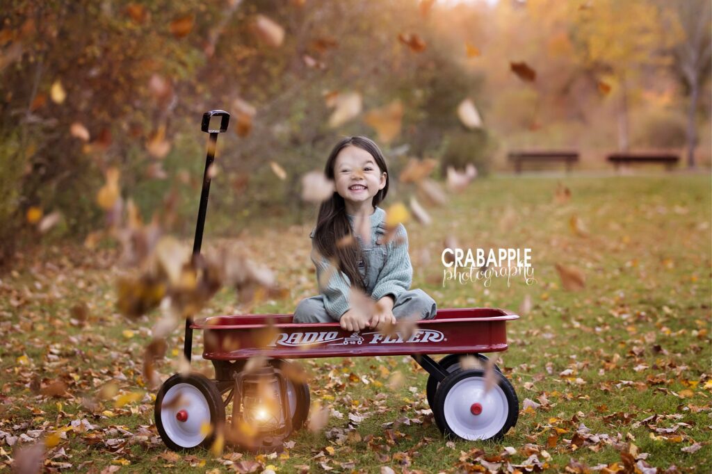 Fun fall in New England outdoor photos with leaves falling and a young girl sitting in a red Radio Flyer wagon.
