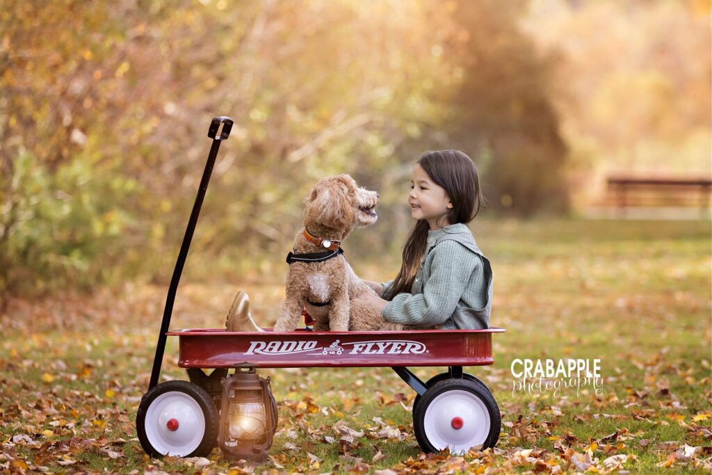 Portrait of a girl and her puppy looking at each other while they sit in a red Radio Flyer wagon.