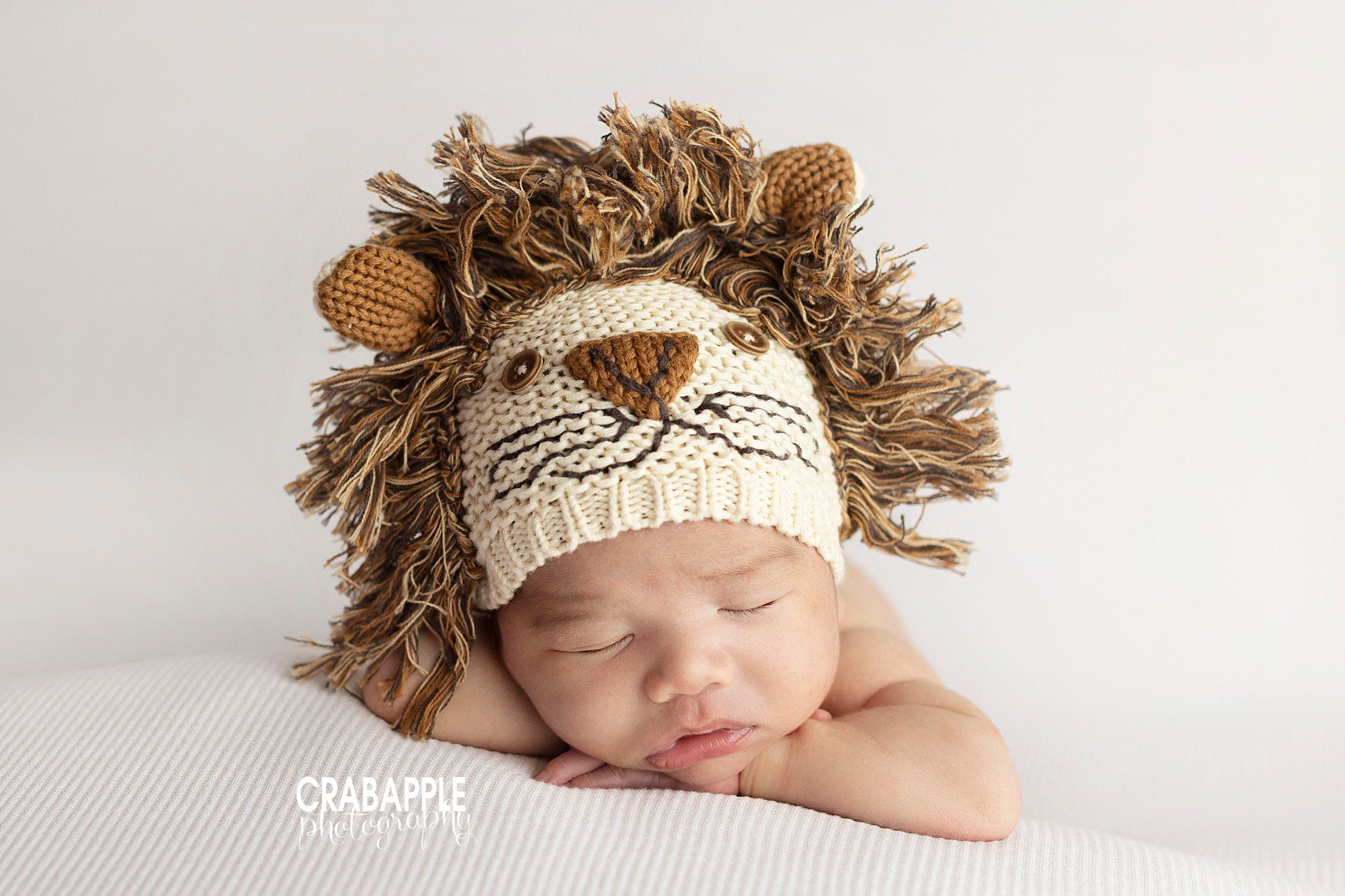 Newborn baby boy wearing a knit lion hat laying on a white blanket.