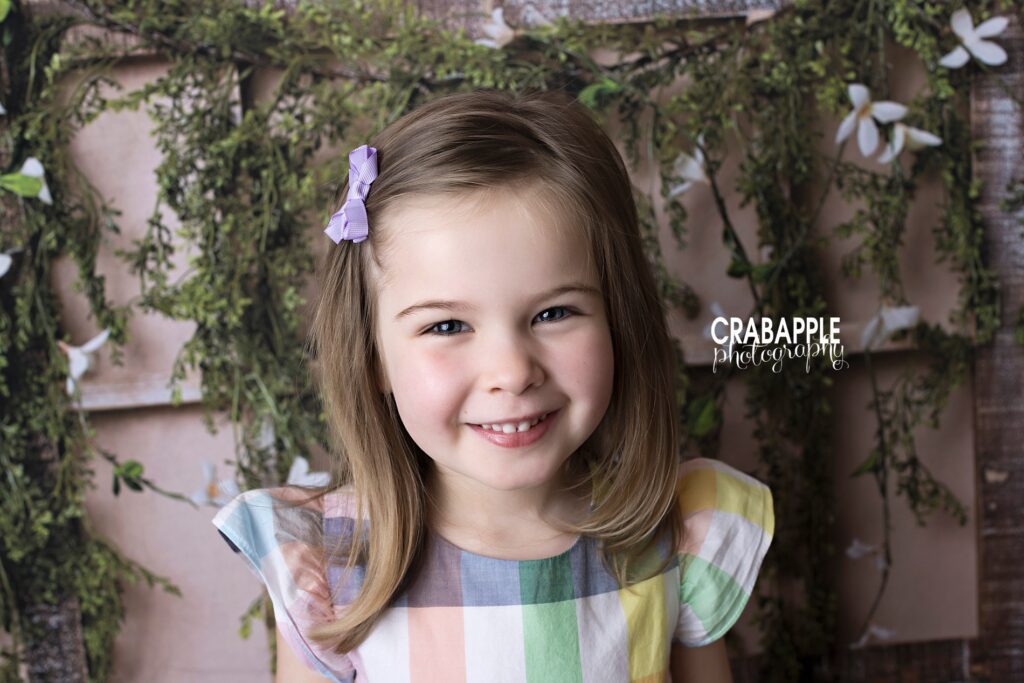 Spring child portrait, close up of a girl wearing a pastel plaid ruffle sleeved dress in front of a rustic backdrop.