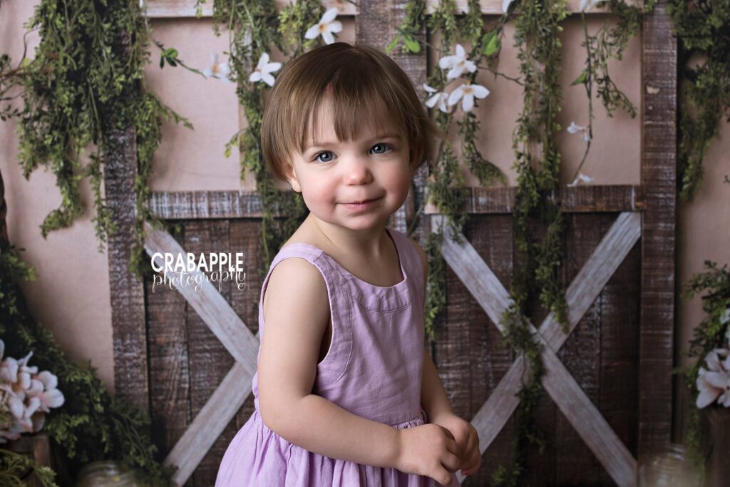 spring themed toddler portraits of a girl in a light purple dress standing in front of a rustic barndoor backdrop with flowers and greenery.