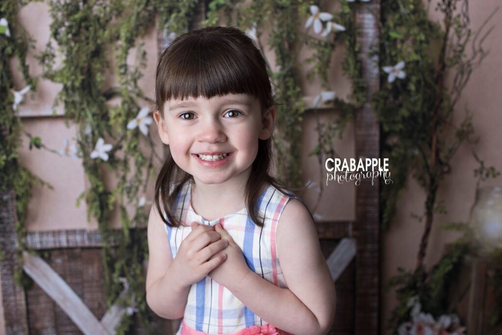 Spring child portrait, close up of a smiling girl wearing a pastel plaid dress in front of a rustic backdrop.