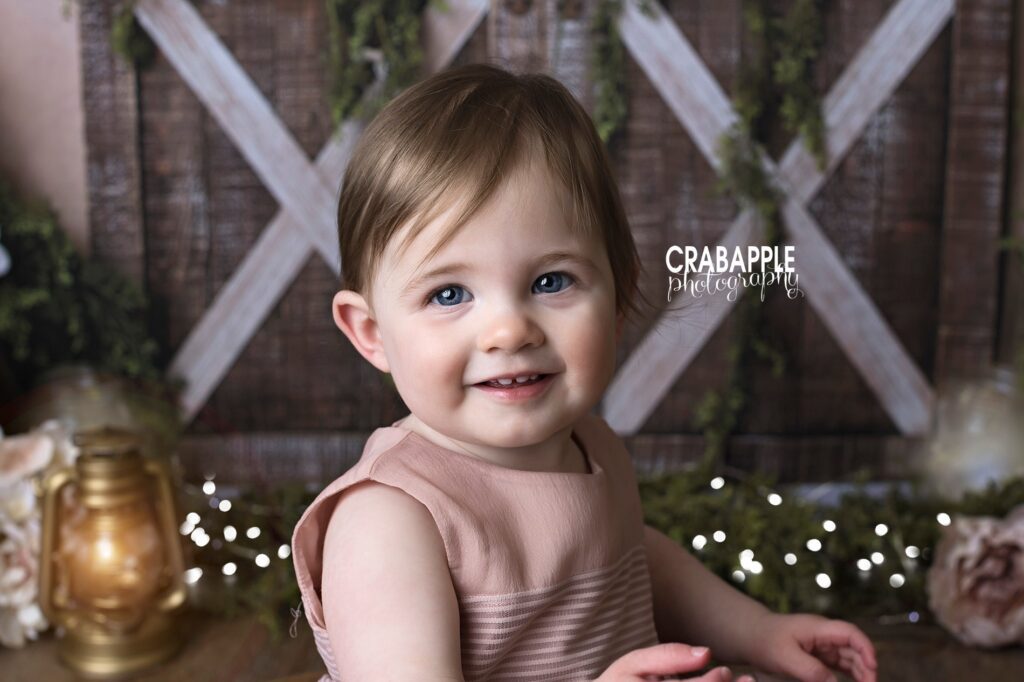 Close up photo of a baby girl smiling at the camera and wearing a light pink dress. The background is a rustic barn door set design.