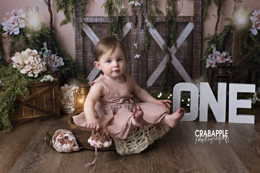 Photo of a one year old baby girl smiling at the camera and wearing a light pink dress. The background is a rustic barn door set design. There are wooden letters spelling the word one.