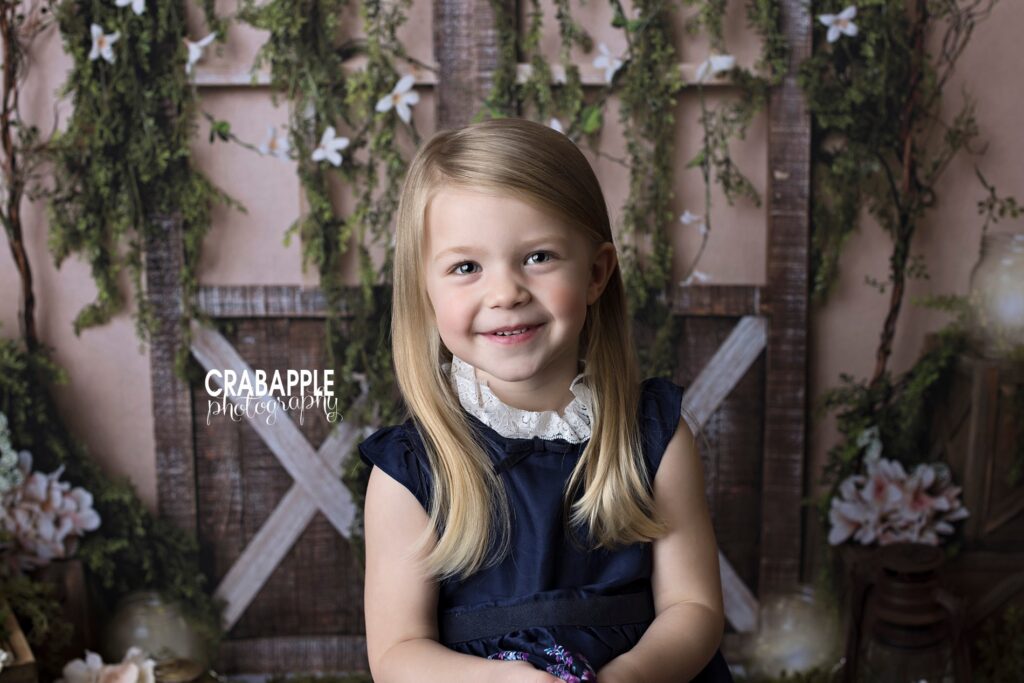 Close up of a young girl wearing a navy blue dress and smiling at the camera in front of a boho backdrop with barn doors and flowers.