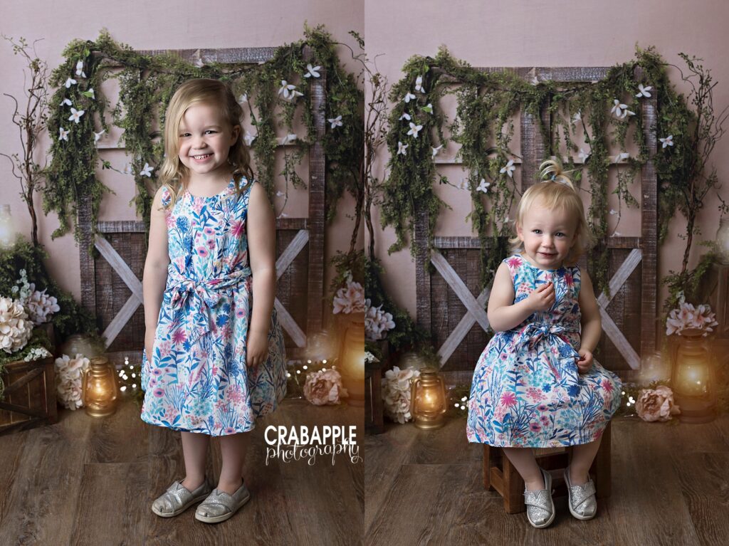 Portraits of two sisters wearing matching floral dresses. The older sister is standing while the younger sister is sitting. The background is a cottagecore barn door setting.