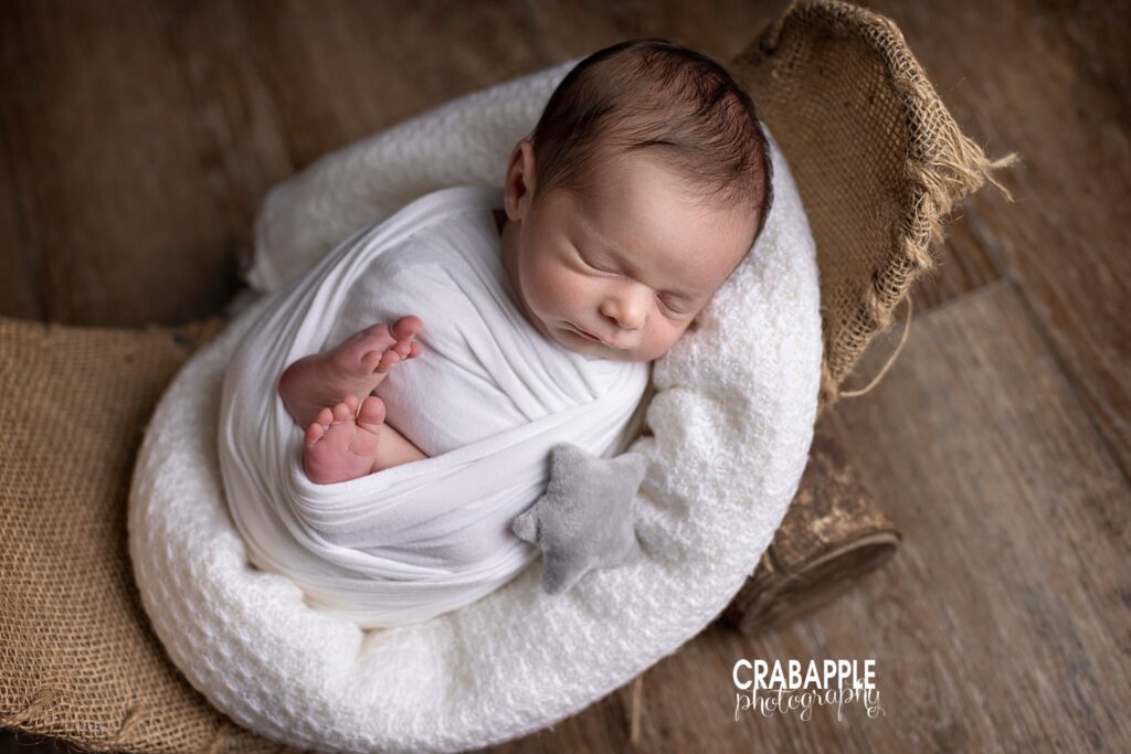 Newborn baby boy swaddled in white with a small gray stuffed star. He is on a burlap prop.