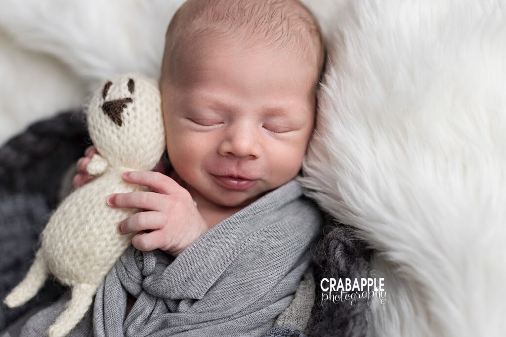 Newborn baby boy slightly smiling as he holds a knit toy. He is wrapped in gray and on a white fur.