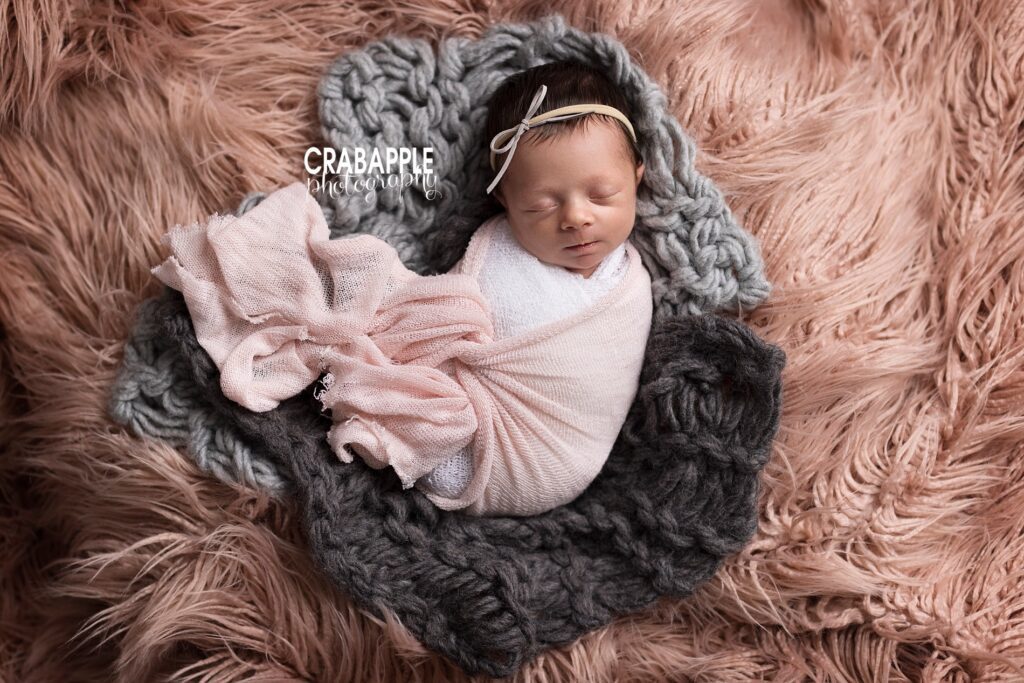 Sleeping newborn portrait of a baby girl wrapped in white and pink swaddles nestled on a chunky knit gray blanket on top of dusty pink fur.