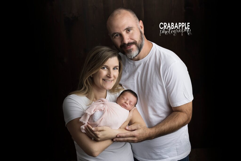 Family portrait with mom and dad wearing white tee shirts in front of a black background. Mom is holding newborn daughter who is swaddled in pink.