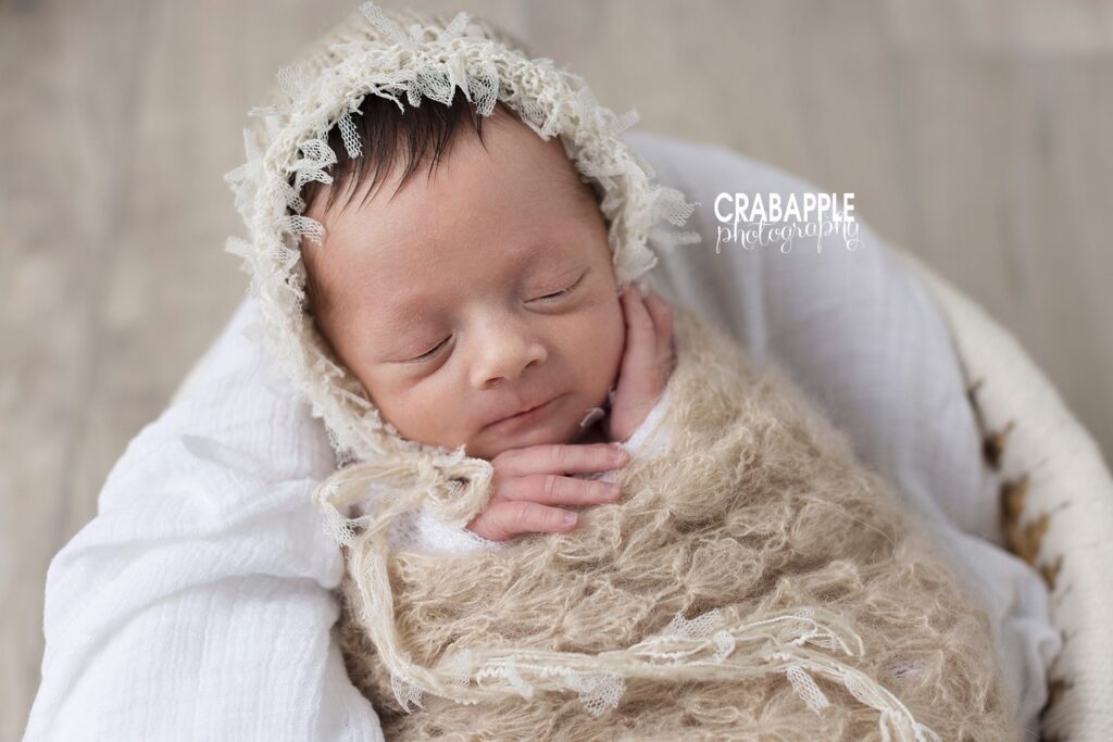 Vintage inspired neutral newborn portrait of a sleeping baby girl wrapped in a beige knit wrap and nestled on a white blanket. She is wearing a knit bonnet with lace details.